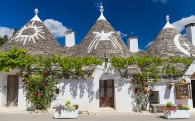Trulli con simboli sui tetti, circondati da fiori e verde. Un'idea per scoprire **paesi da visitare vicino Alberobello**.