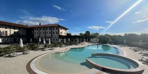 Piscina a forma di rene con idromassaggio annesso, circondata da lettini e ombrelloni, di fronte a un hotel in stile piemontese.