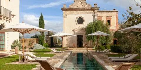 Piscina e lettini in una masseria per bambini in Puglia, con un antico edificio sullo sfondo.