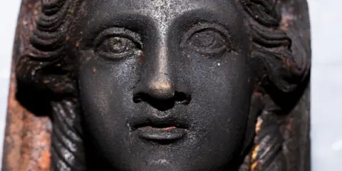 Volto di bronzo scuro, un dettaglio dei bronzi di San Casciano dei Bagni, con capelli a onde e trecce.