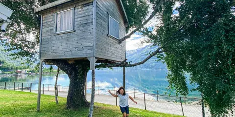 Bambina corre verso una casa sull'albero in legno, con un lago e montagne sullo sfondo.