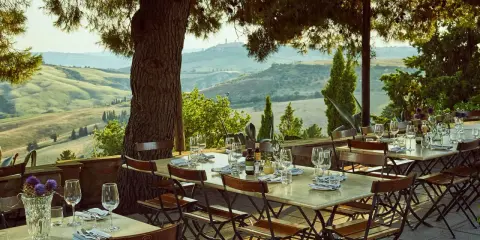 Tavoli apparecchiati sotto un pergolato con vista sulle dolci colline dell'entroterra toscana. Un invito a gustare la cucina locale.