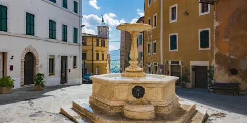 Fontana in pietra nella Valdichiana Senese, circondata da edifici storici con persiane verdi e un cielo azzurro con nuvole bianche.