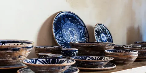 Ceramiche artigianato toscano con decori blu su fondo grezzo. Piatti e ciotole disposti su una mensola di legno.