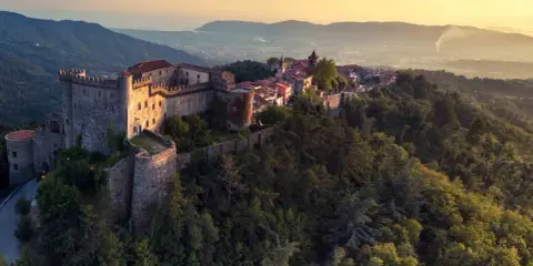 Antico castello medievale su una collina boscosa, uno dei tanti castelli da visitare in Toscana, con un villaggio arroccato e vista sulla valle al tramonto.