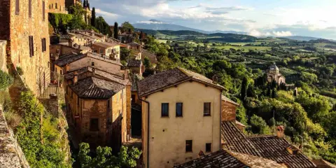 Panoramica di borghi toscana in treno, con case in pietra e tegole rosse su colline verdi sotto un cielo nuvoloso.