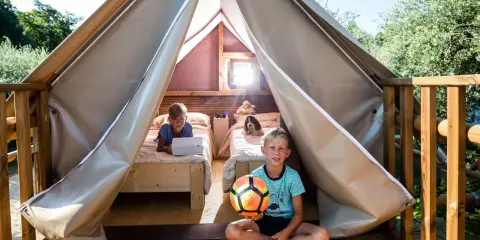 Bambini felici in un glamping: uno gioca col pallone, l'altro usa un tablet a letto. Avventura e relax per i più piccoli.