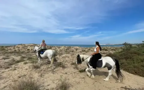 Due donne godono di una passeggiata a cavallo in Sicilia, tra dune sabbiose e il mare azzurro.