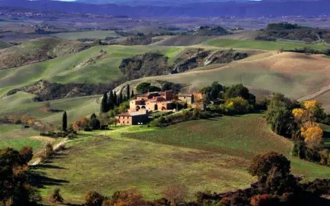 Colline ondulate e campi coltivati, un casale in mattoni rossi e cipressi svettanti, tipici delle **strade panoramiche toscana**.