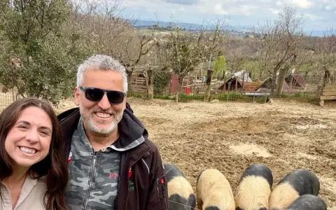 Tre amici sorridono mentre salutano i maiali senesi in un recinto fangoso, con un paesaggio collinare sullo sfondo.