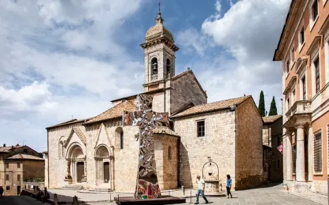 Piazza a San Quirico con una scultura moderna a forma di croce riflettente davanti alla chiesa romanica.
