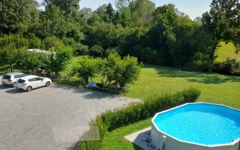 Un **agricampeggio con piscina** immerso nel verde. Auto parcheggiate, camper, biciclette e persone che si rilassano all'ombra degli alberi. La piscina azzurra invita a un tuffo rinfrescante.