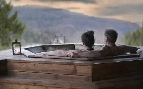 Coppia abbracciata in una vasca idromassaggio di legno, con lanterne accese e vista sulle montagne. Un momento di puro relax in Trentino.