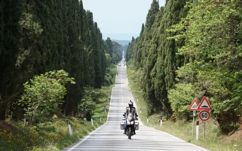 Un motociclista percorre un viale cipressi toscana, immerso nel verde e con il mare all'orizzonte.