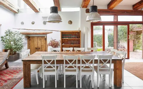 Un rustico tavolo in legno con sedie bianche, illuminato da lampade industriali, in una casa con travi a vista e un giardino che ricorda il Val di Cecina.