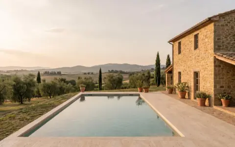 Piscina con vista sulle dolci colline pisane al tramonto. Una casa in pietra e ulivi completano il paesaggio idilliaco.