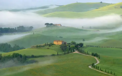 Dolci colline verdi avvolte dalla nebbia mattutina nel Val di Norcia, con casali in pietra e filari di cipressi.