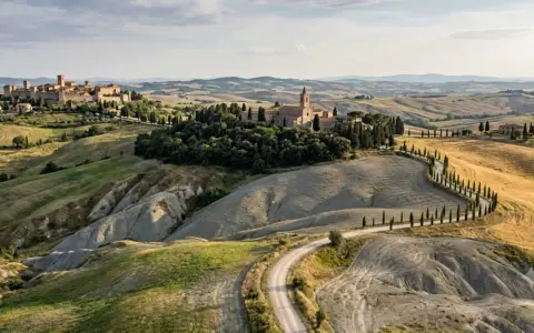 Dolci colline senesi con un borgo medievale e un'abbazia, attraversate da una strada sinuosa fiancheggiata da cipressi.
