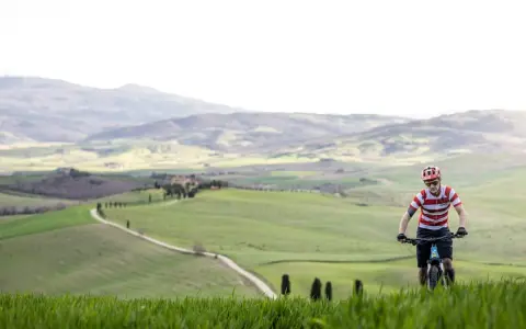 Ciclista in un paesaggio collinare della Toscana, con un sentiero tortuoso che serpeggia tra campi verdi.