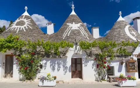 Trulli con simboli sui tetti, circondati da fiori e verde. Un'idea per scoprire **paesi da visitare vicino Alberobello**.