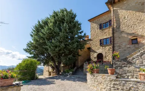 Un casale in pietra con un grande cipresso, tipico degli agriturismi del centro Italia, circondato da fiori in vaso e con un panorama collinare.