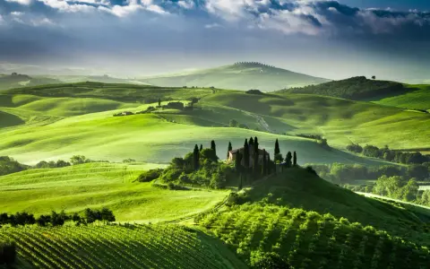 Colline verdi ondulate del Val d'Orcia, con una casa colonica e cipressi su una collina. Un paesaggio da sogno, quasi una mappa visiva della Toscana.