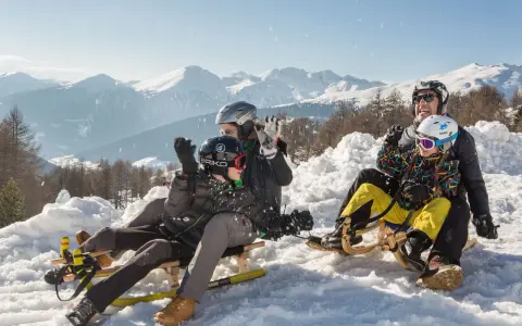 Famiglia felice in slittino sulla montagna per bambini in estate, con neve e montagne innevate sullo sfondo.