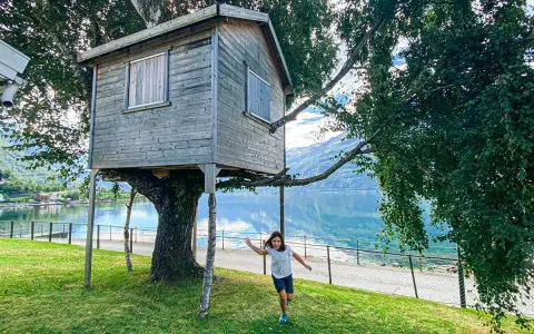 Bambina corre verso una casa sull'albero in legno, con un lago e montagne sullo sfondo.