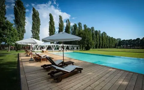 Una splendida piscina in campagna, con lettini prendisole in legno e ombrelloni bianchi, circondata da un prato verde e alti alberi.