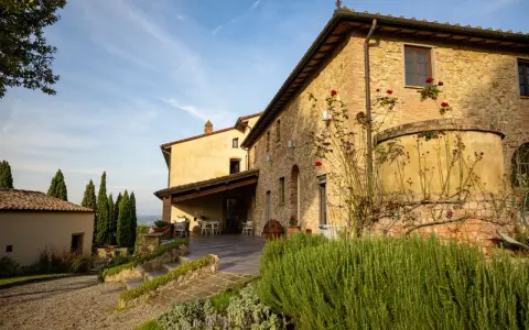 Un casale in pietra con rose rampicanti, un pergolato con tavoli e sedie, e cipressi svettano nel paesaggio della **Toscana in inverno**.