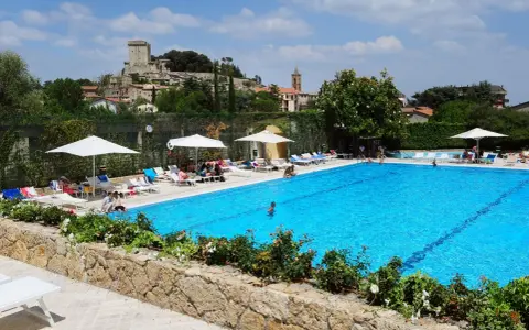 Piscina azzurra in un villaggio famiglie toscana, con castello medievale sullo sfondo e persone che si rilassano al sole.