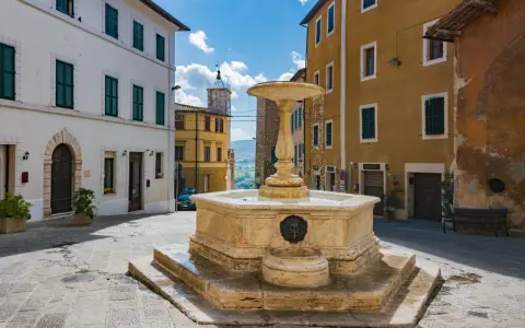 Fontana in pietra nella Valdichiana Senese, circondata da edifici storici con persiane verdi e un cielo azzurro con nuvole bianche.