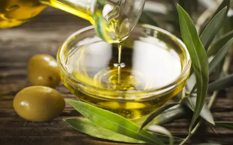 Olio di oliva non extravergine versato in una ciotola di vetro, con olive e foglie d'ulivo su fondo di legno.