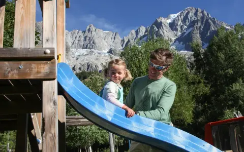 Un padre e una bambina si divertono su uno scivolo in un parco giochi con le maestose montagne della Valle d'Aosta sullo sfondo, un'estate indimenticabile.