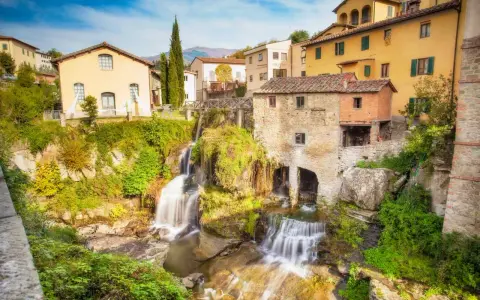 Cascata e mulino in pietra, tra i borghi della Toscana. Case gialle e verdi colline sullo sfondo.