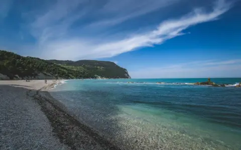 Spiaggia di ciottoli con mare cristallino e una collina verdeggiante, tipica del entroterra marchigiano.