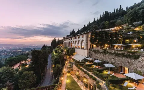 Un lussuoso hotel si affaccia sulle **colline di Firenze** al tramonto, con giardini illuminati e la città in lontananza.