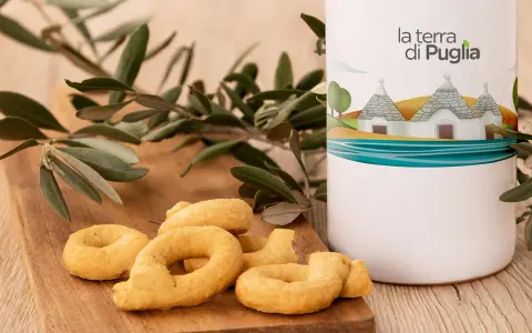 Taralli pugliesi su tagliere di legno, accanto a un contenitore con il logo "la terra di Puglia" e un ramo d'ulivo.