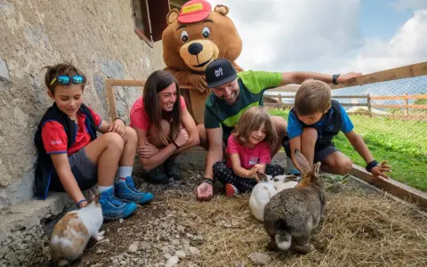 Bambini e adulti interagiscono con conigli in un recinto, parte di un'esperienza in fattorie didattiche. Un personaggio con costume da orso sorride.