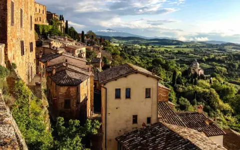 Panoramica di borghi toscana in treno, con case in pietra e tegole rosse su colline verdi sotto un cielo nuvoloso.