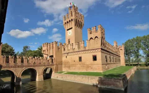 Un imponente castello in mattoni rossi, uno dei tanti castelli Emilia Romagna, con un ponte ad archi sull'acqua e torri merlate sotto un cielo azzurro.