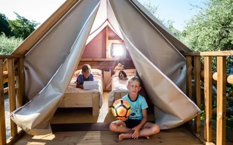 Bambini felici in un glamping: uno gioca col pallone, l'altro usa un tablet a letto. Avventura e relax per i più piccoli.