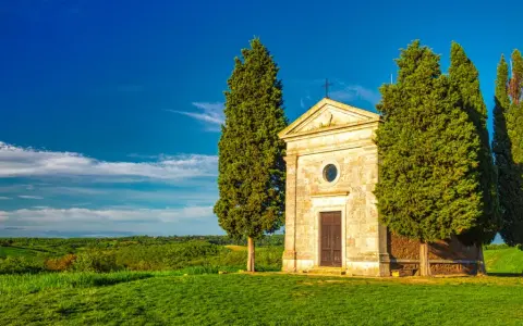Cappella di San Quirico d'Orcia, circondata da cipressi, su un prato verde sotto un cielo azzurro.