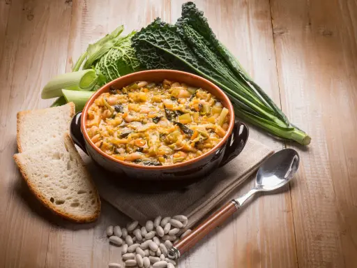Piatto tipico toscano con pane raffermo: ribollita fumante in ciotola, contornata da fagioli, cavolo nero e porri.