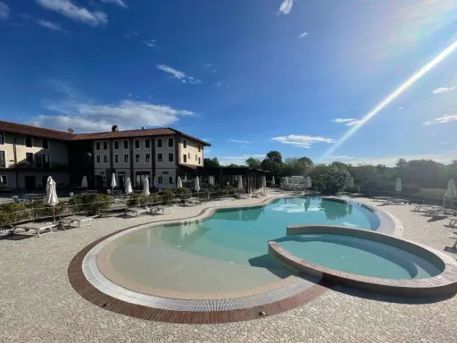 Piscina a forma di rene con idromassaggio annesso, circondata da lettini e ombrelloni, di fronte a un hotel in stile piemontese.