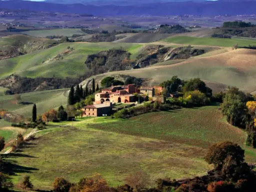 Colline ondulate e campi coltivati, un casale in mattoni rossi e cipressi svettanti, tipici delle **strade panoramiche toscana**.