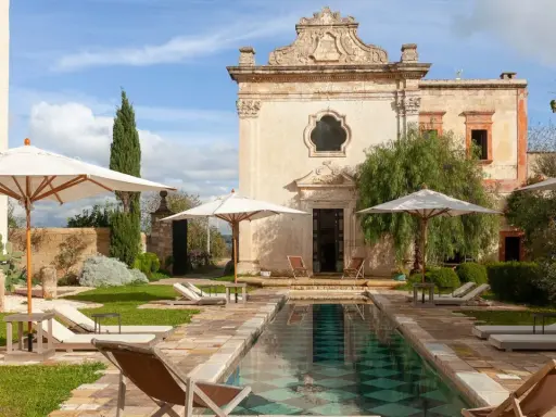 Piscina e lettini in una masseria per bambini in Puglia, con un antico edificio sullo sfondo.