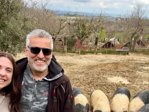 Tre amici sorridono mentre salutano i maiali senesi in un recinto fangoso, con un paesaggio collinare sullo sfondo.