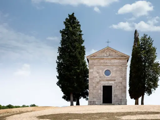 La cappella della Madonna di Vitaleta, con i suoi cipressi iconici, si erge solitaria su una collina toscana sotto un cielo azzurro e nuvoloso.