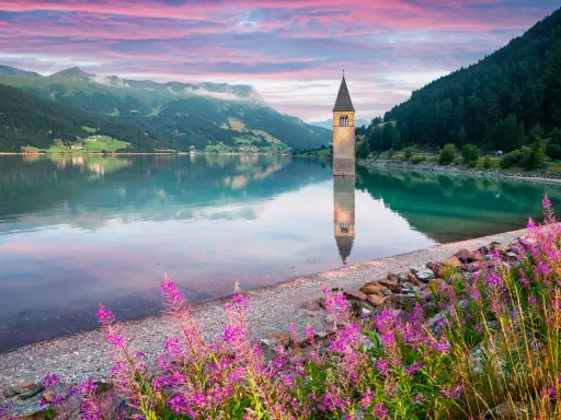 Torre sommersa in un lago alpino, con montagne verdi e cielo rosa all'alba. Un angolo di montagna Friuli Venezia Giulia incantevole.